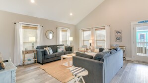 A spacious living room with a grey sofa set, a coffee table, and a small wooden side table. There are large windows with white curtains and a view of the outside.