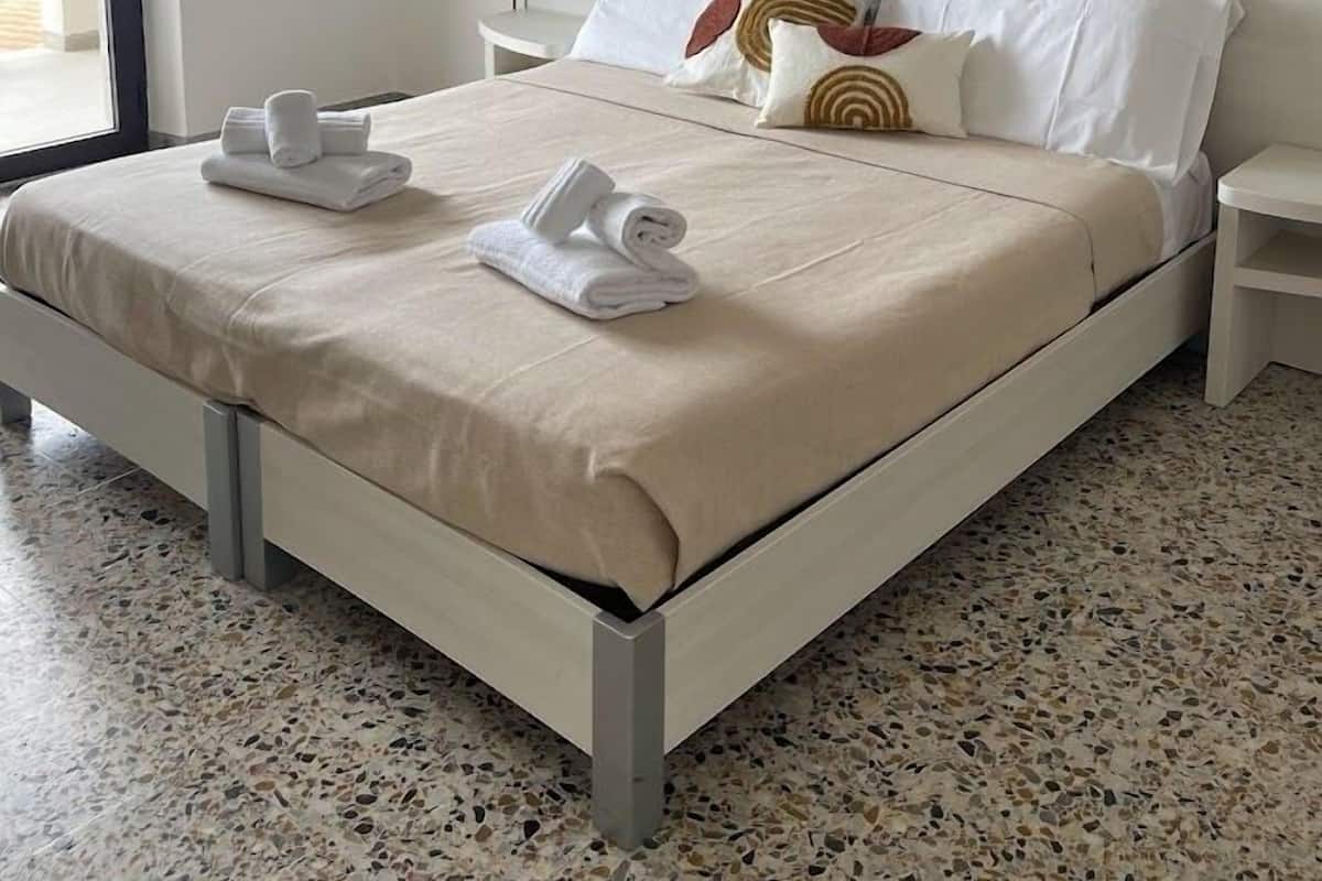 A neatly made bed with white linens and towels, a headboard with a decorative piece, and a simple nightstand with a lamp.