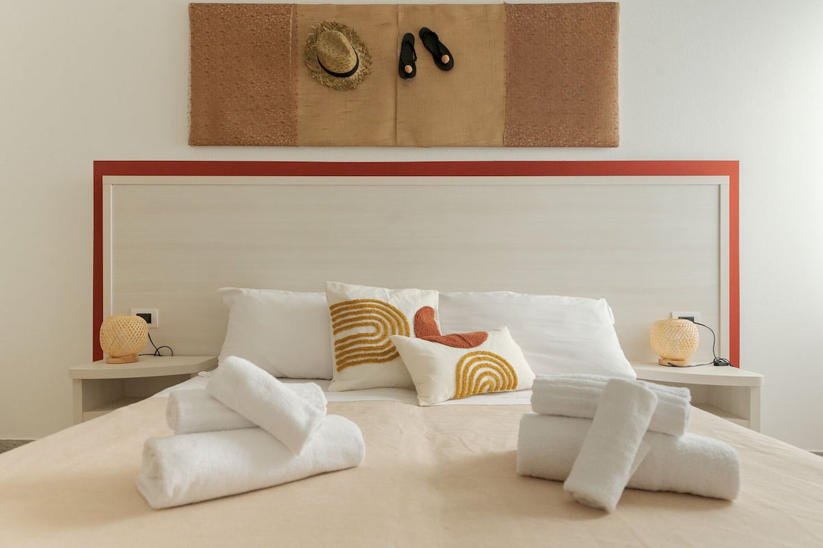 A neatly made bed with white linens, two decorative pillows, and a pair of slippers on a wall-mounted board.
