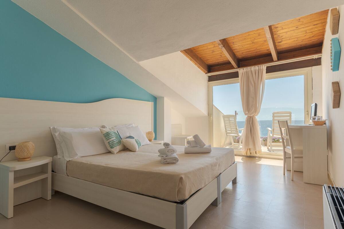 A bedroom with a bed, nightstands, a desk, and a view of the sea.