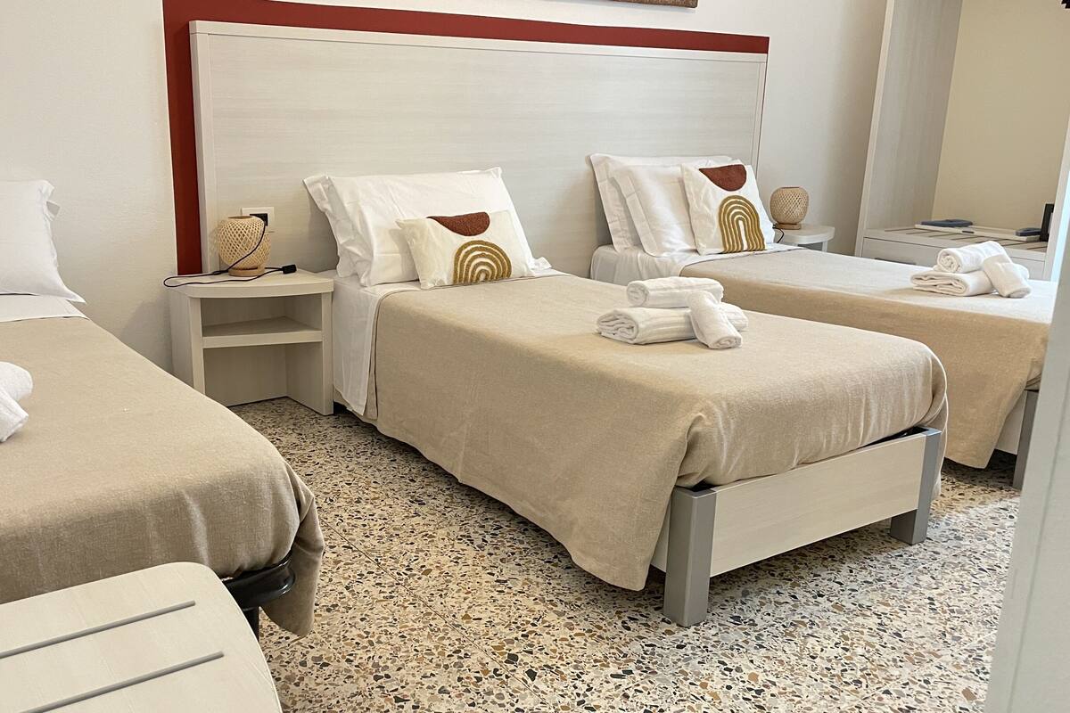A hotel room with two beds, each with a beige bedspread and pillows, a nightstand with a lamp, and a small table with a rolled towel.