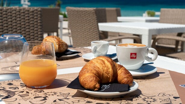 A table with croissants, a glass of orange juice, and a cup of coffee, set against a backdrop of a beach and ocean view. - Hotel Maluentu (San Vero Milis)