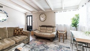A living room with a beige sofa, a leather recliner, a glass coffee table, and a dining table with chairs. A large wall clock is visible, and a door is seen in the background. - The Perfect Vacation Spot Near Six Flags (Santa Clarita)