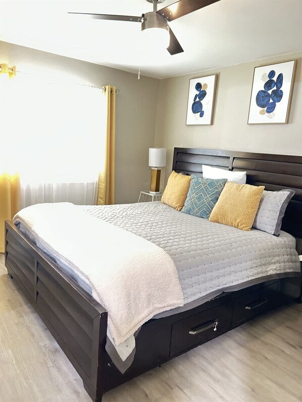 A bedroom with a dark wooden bed, a white quilted bedspread, and multiple pillows. There is a ceiling fan, a bedside table with a lamp, and artwork on the wall.