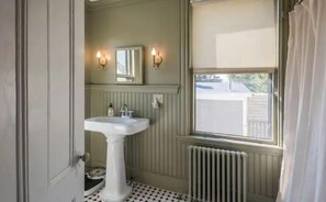 Une salle de bain avec un lavabo sur colonne, un miroir, deux appliques murales, un radiateur et une fenêtre donnant sur l’extérieur.