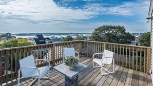 Een houten terras met witte metalen stoelen en een tafel, met uitzicht op een stadsgezicht en water.