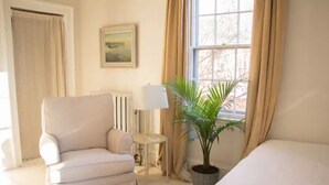 Una habitación con cama, una silla, una lámpara, un radiador, una ventana con cortinas y una planta en maceta.