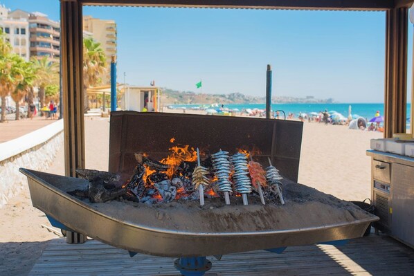 A beachside grill with skewers of food over an open flame, overlooking a sandy beach with umbrellas and palm trees.