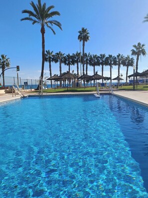A clear blue pool with palm trees and umbrellas in the background.