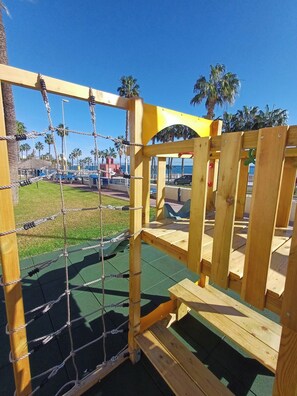 A wooden play structure with a rope net, surrounded by a green surface, with palm trees and a clear sky in the background.