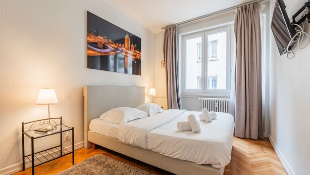 A neatly arranged bedroom with a bed, two bedside lamps, a nightstand, a window with curtains, and a cityscape painting on the wall. - Le Kléber  - Appt en hypercentre, proche Place Kléber (Strasbourg)