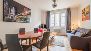 A modern living room with a dining area, a sofa, a TV, and a large window with curtains. - Le Kléber  - Appt en hypercentre, proche Place Kléber (Strasbourg)