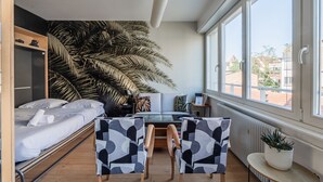 Un dormitorio compacto con cama, escritorio, dos sillas, un ventanal amplio y una planta en maceta.