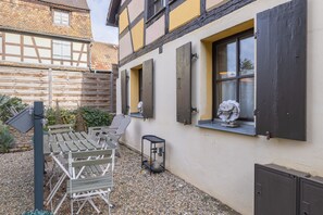 Une cour avec une table et des chaises en bois, un petit jardin et un bâtiment doté d’une façade en pierre et de volets en bois.