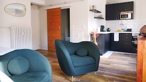 A modern living room with a teal armchair, a wooden floor, and a kitchen area with dark cabinets and a microwave. - Le Cocon Urbain - Joli Studio récemment rénové (Strasbourg)