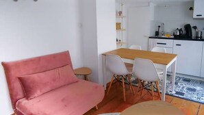 Un espace de vie compact comprenant un canapé rose, une table à manger en bois avec quatre chaises, un coin cuisine et une petite table ronde.