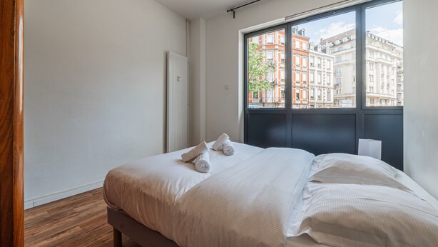 A hotel room with a large window, a bed with white linens, and a view of buildings outside.
