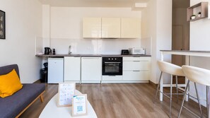Eine moderne Küche mit weißen Schränken, einem schwarzen Backofen und einer Mikrowelle. Es gibt einen kleinen Essbereich mit zwei weißen Barhocker und einem Tisch.