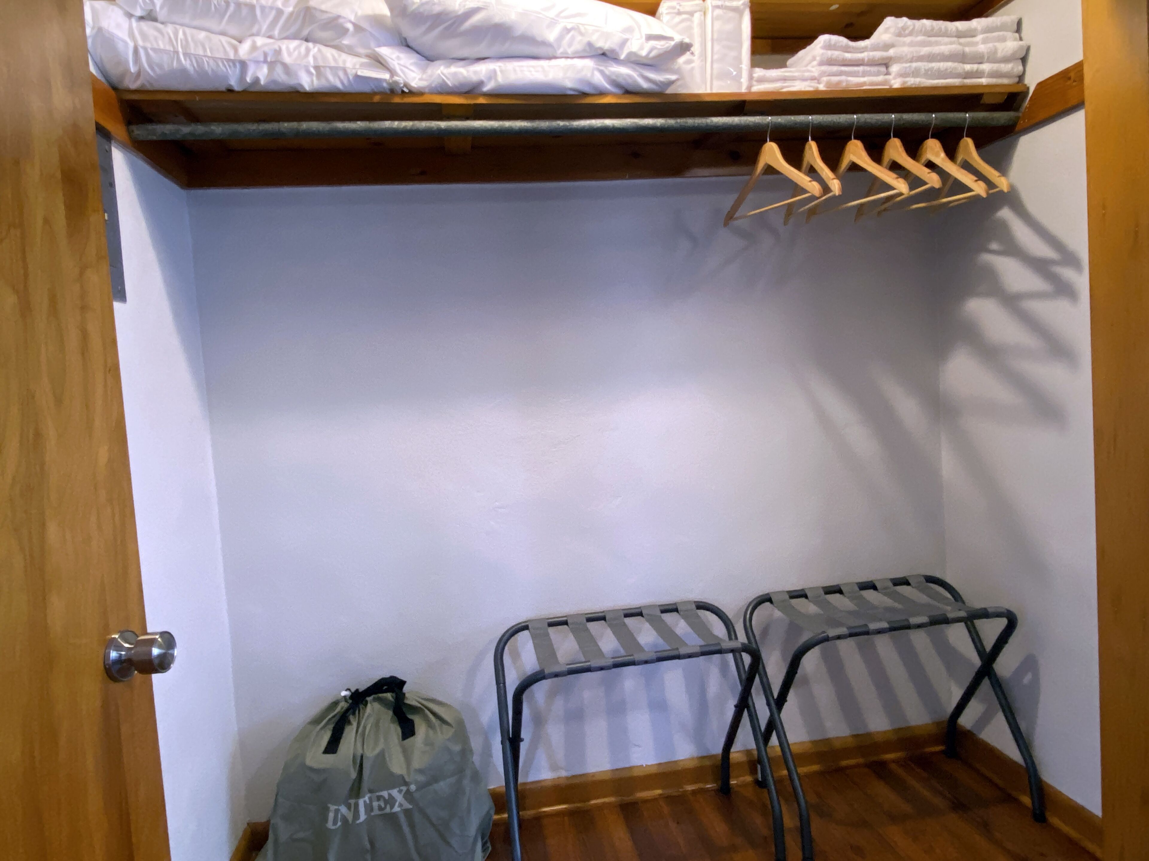 Habitació petita amb un llit, una estendal metàl·lic, un tamboret petit i una bossa a terra.