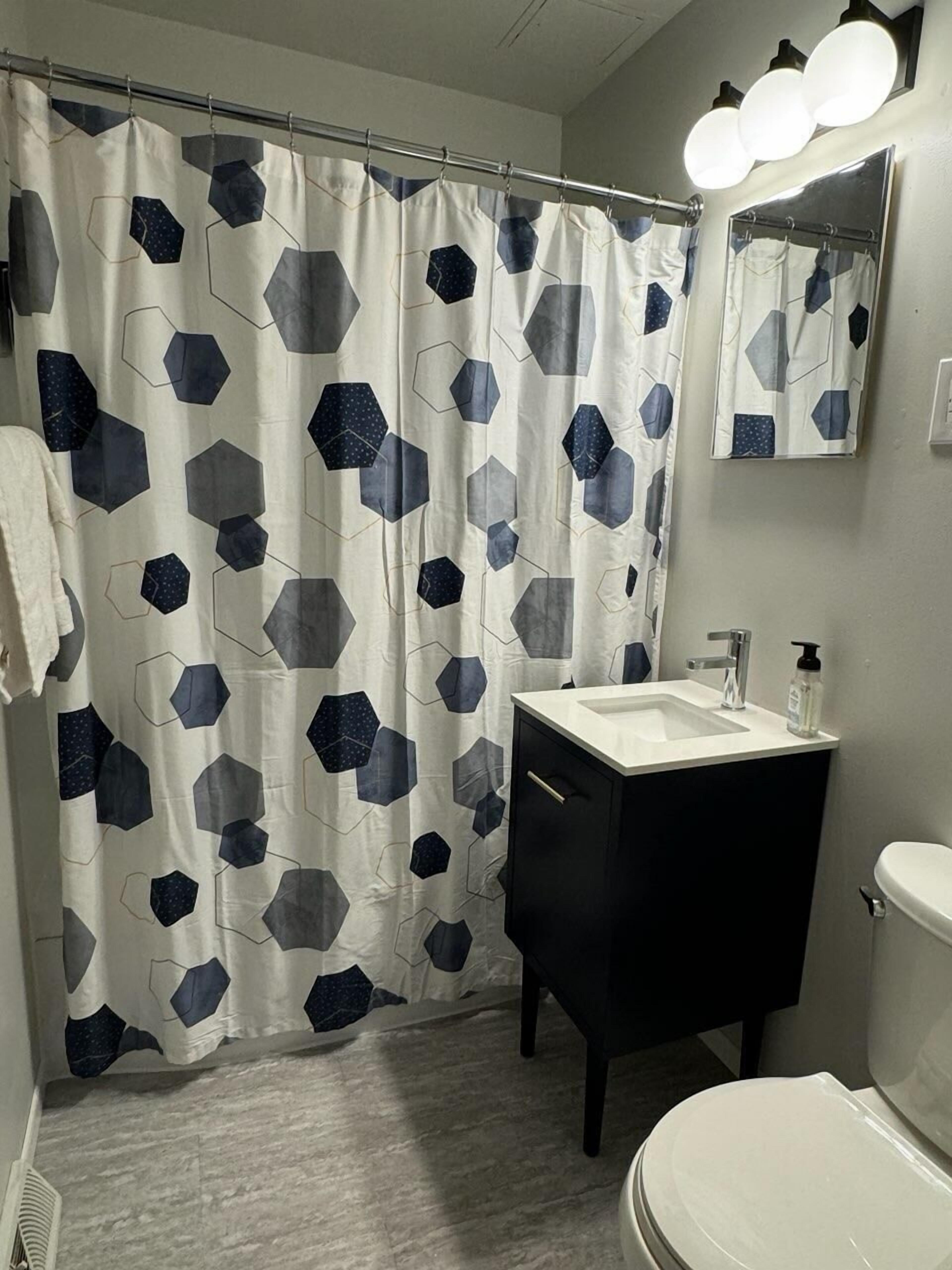 A bathroom with a shower curtain featuring a geometric pattern, a white sink, a black cabinet, a mirror, and a toilet.