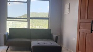 A room with a sofa and ottoman, a wooden door, and a window with a view of hills. - Nos Soño, Condo 3-bdr - Stunning View - Valley Verde 9, St. Maarten 2-4 Guests
 (Philipsburg)