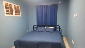 A small, single-bed room with a blue bedspread, a window with blinds, and a curtain. - Nos Soño, Condo 3-bdr - Stunning View - Valley Verde 9, St. Maarten 2-4 Guests
 (Philipsburg)
