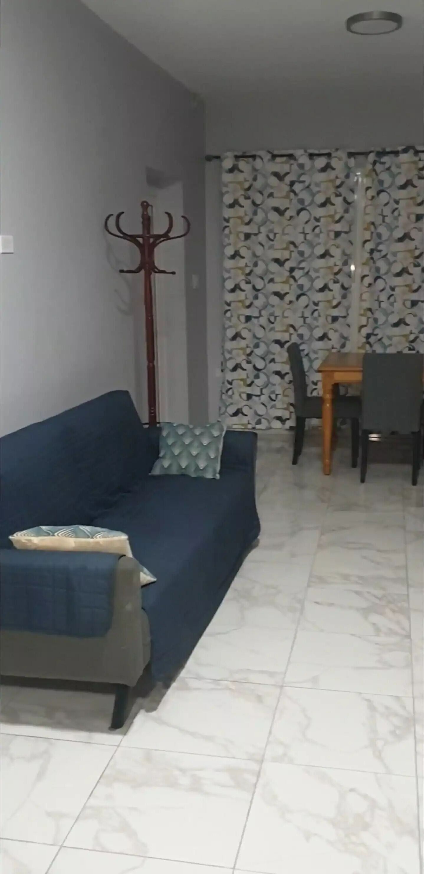 A living room with a blue sofa, a wooden coat rack, a patterned wall, and a dining area with a table and chairs.
