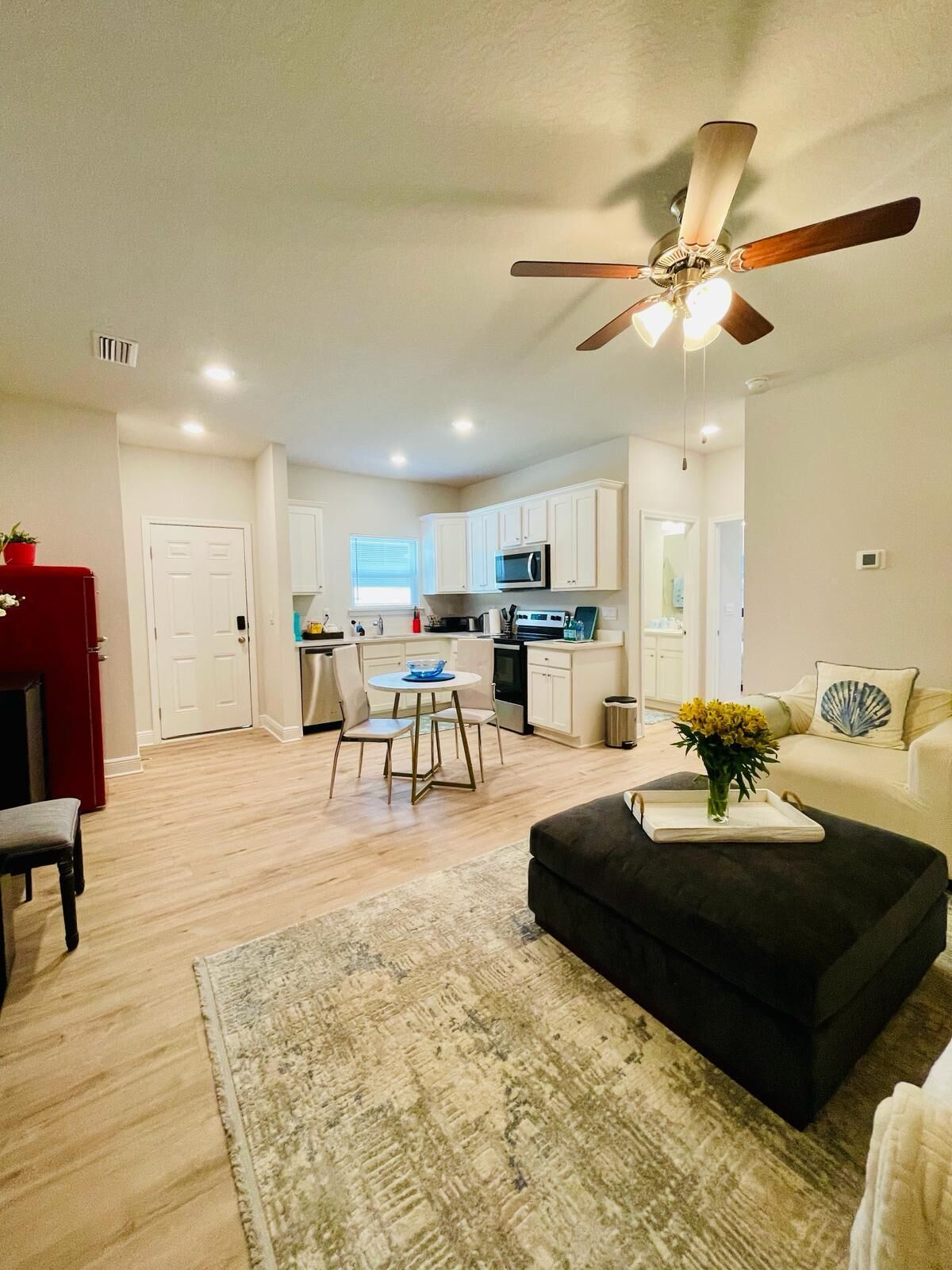 A modern living room with a black ottoman, a dining area with a round table, and a kitchen with white cabinets and a ceiling fan.