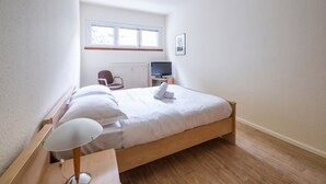A bedroom with a wooden bed, a desk, a chair, a TV, and a window.