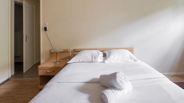A hotel room with a bed, bedside table, lamp, and a clock on the wall.