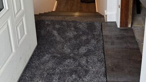 A hallway with a wooden floor, a black rug at the entrance, a door with a keyhole, a wall-mounted sign, and a small shelf with a vase of flowers.