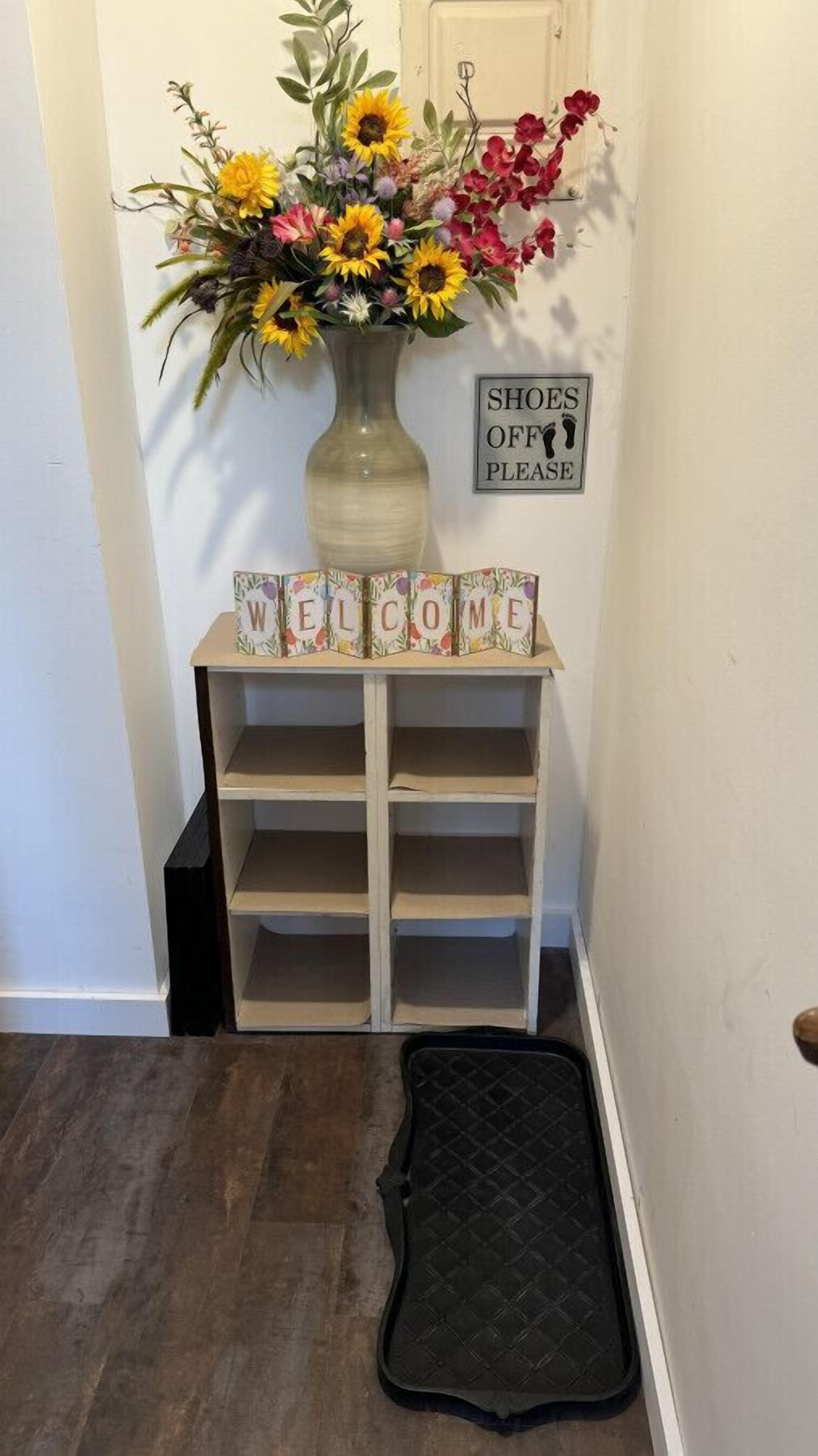Une pancarte d’accueil avec le mot « BIENVENUE », un vase rempli de fleurs et une affiche sur le mur indiquant : « VEUILLEZ ENLEVER VOS CHAUSSURES ».