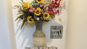 A welcome sign with 'WELCOME' letters, a vase with flowers, and a 'SHOES OFF PLEASE' sign on a wall.