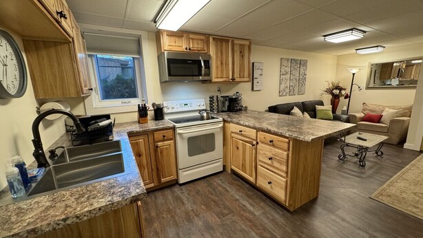 A kitchen with wooden cabinets, a stainless steel sink, a microwave, and a stove. There is a window above the sink and a clock on the wall.