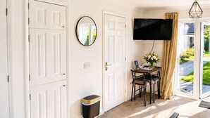 A room with white doors, a flat-screen TV, a small table with a vase of flowers, and a view of a garden through a glass door. - Lodge 6 - Lytton Tree Lodge - Reydon (Reydon)