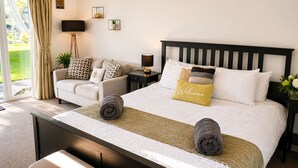 A bedroom with a bed, a sofa, a TV, a lamp, and a window with curtains. - Lodge 6 - Lytton Tree Lodge - Reydon (Reydon)