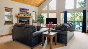 Ein geräumiges Wohnzimmer mit Kamin, zwei Sofas, einem Couchtisch und großen Fenstern mit Blick ins Freie.
