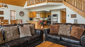 Ein großzügiger Wohnbereich mit hohen Decken und einer Holztreppe. Im Hintergrund ist eine Küche mit Holzausstattung und modernen Geräten zu sehen.