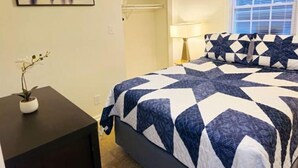 Un dormitorio con cama, mesita de noche, un cuadro, planta en maceta y ventana con persianas.