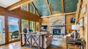 A spacious living room with a stone fireplace, wooden ceiling beams, and a large window overlooking a scenic view. - Dream Catcher Mountain View Cabin (Pigeon Forge)