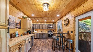 A wooden interior with a kitchen area, bar stools, and a large window with a view of the outdoors. - Ultimate Smoky Getaway: Hot Tub, Fun & Fresh Air in the Heart of the Smokies. (Sevierville)