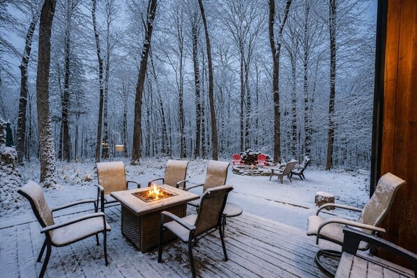 An outdoor wooden deck with a fire pit and chairs, surrounded by a snowy forest.