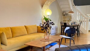 A modern living room with a yellow sofa, a wooden coffee table, and a blue leather ottoman. There is a dining area with a table and chairs, and a staircase in the background. - Le Bijou du Minage - Jardin/terrasse - Hypercentre LA Rochelle (La Rochelle)