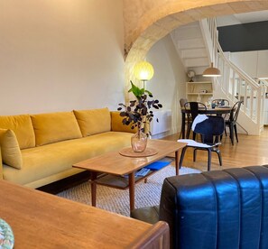 A modern living room with a yellow sofa, a wooden coffee table, and a blue leather ottoman. There is a dining area with a table and chairs, and a staircase in the background. - Le Bijou du Minage - Jardin/terrasse - Hypercentre LA Rochelle (La Rochelle)
