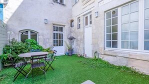 Un patio con una pequeña mesa y sillas, rodeado de césped cuidado y plantas en macetas.
