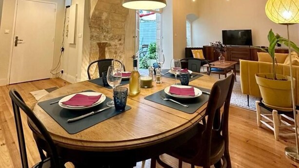 Un comedor con arco de piedra, mesa de madera y sillas. Se observa un televisor y una silla amarilla al fondo.