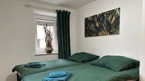 Un dormitorio con cama, ventana con cortinas, un florero con plantas y un cuadro en la pared.
