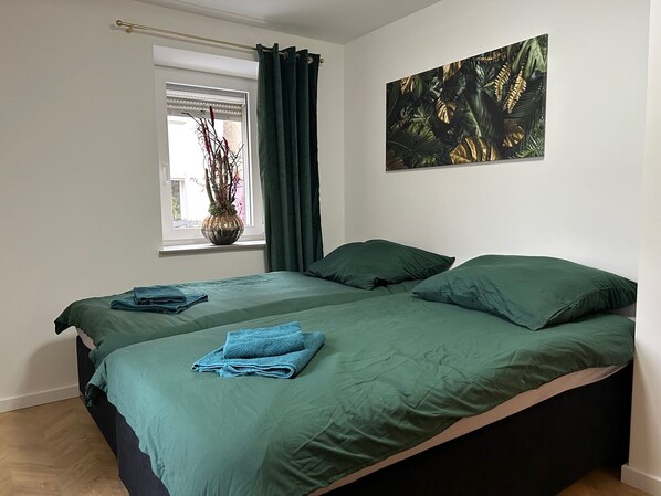 A bedroom with a bed, a window with curtains, a vase with plants, and a painting on the wall.