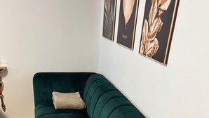 Un sofá color turquesa con un cojín, una mesita de noche con estampado dorado y verde, e impresiones botánicas enmarcadas en la pared.