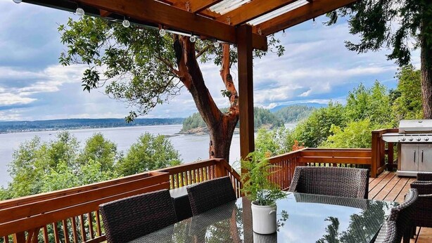 A covered outdoor deck with a glass-top table and wicker chairs, overlooking a lake.